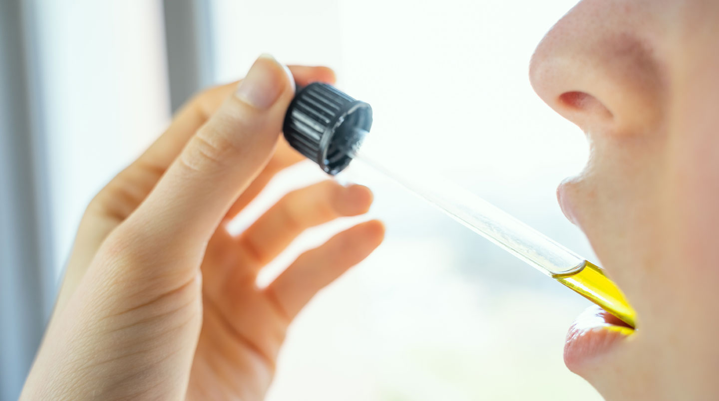 Gros plan macro d'une goutte dorée translucide d'huile CBD suspendue à l'extrémité d'un compte-gouttes en verre, arrière-plan flouté aux teintes chaudes