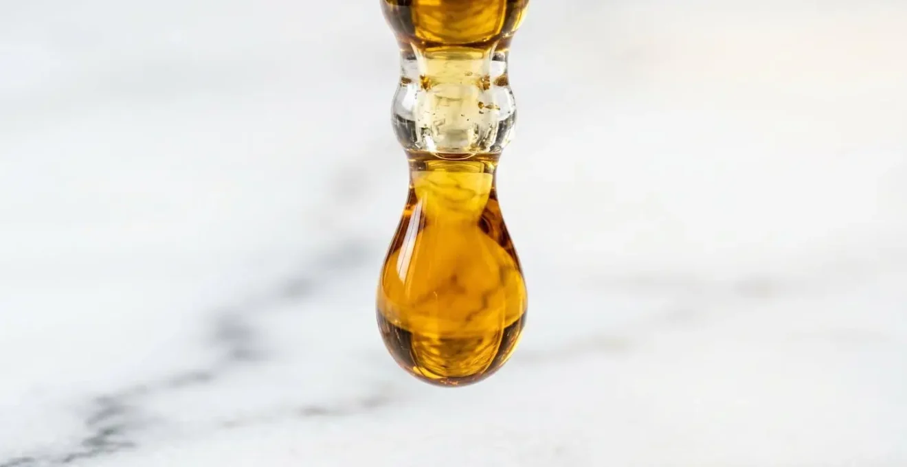 Gros plan macro d'une goutte dorée translucide d'huile CBD suspendue à l'extrémité d'un compte-gouttes en verre, arrière-plan flouté aux teintes chaudes